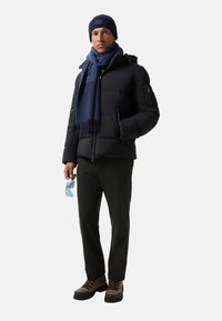 Man in een zwarte pufferjack, marineblauwe beanie, blauwe sjaal, antracietkleurige broek, bruine wandelschoenen, met blauwe zonnebril in de hand.