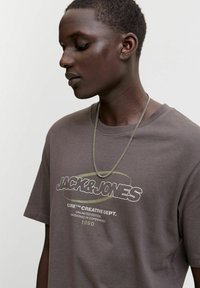 Mlada osoba s kratkom kosom koja nosi sivi Jack & Jones majicu i srebrnu lančić, na jednobojnoj svijetloj pozadini.