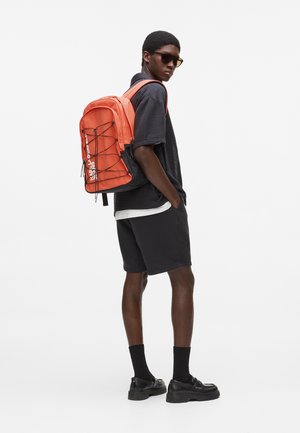 Joven con pantalones cortos y camiseta oscuros, zapatos negros, gafas de sol y llevando una gran mochila naranja con cuerdas elásticas negras.