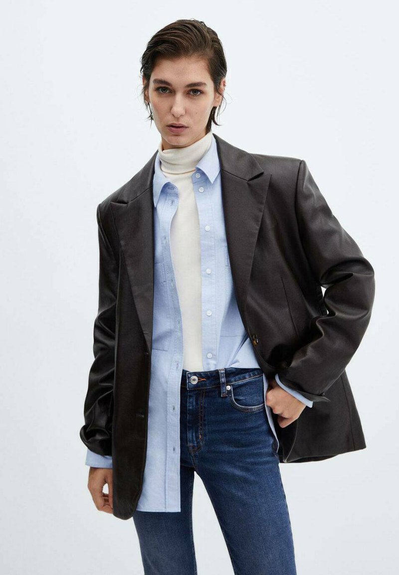 Blazer noir en simili cuir sur une chemise bleu clair à boutons, col roulé blanc en dessous, associé à un jean foncé. Texture lisse, coupe ajustée.