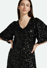Vestido negro de lentejuelas con escote en V, mangas cortas abullonadas y corte holgado, que presenta una superficie brillante y texturizada.