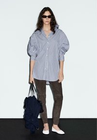 Camicia oversize a righe blu e bianca, pantaloni in suede marrone e scarpe bianche. Borsa nera con un maglione blu navy. Modella con occhiali da sole.