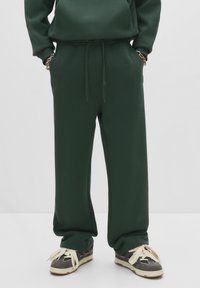 Person in dunkelgrünen Jogginghosen mit Händen in den Taschen und grauen Sneakers mit großen cremefarbenen Schnürsenkeln auf einem weißen Boden.