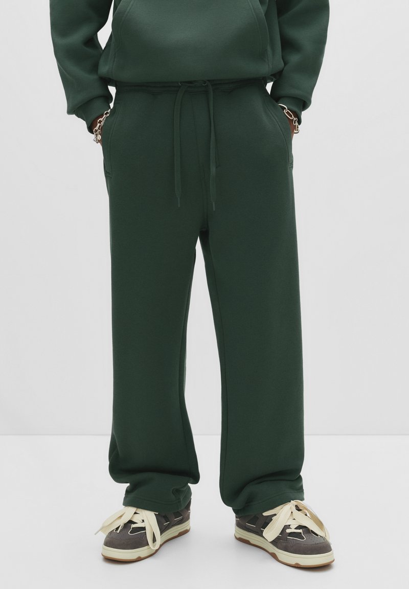 Person in dunkelgrünen Jogginghosen mit Händen in den Taschen und grauen Sneakers mit großen cremefarbenen Schnürsenkeln auf einem weißen Boden.