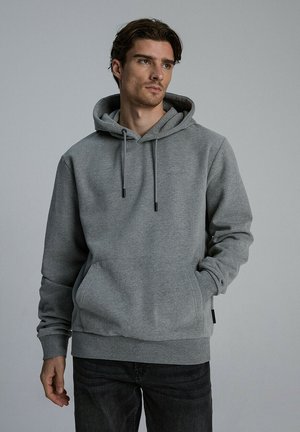 Szara bluza z kapturem, wykonana z miękkiego materiału, z przednią kieszenią, regulowanymi sznurkami oraz ściągaczami u rękawów i u dołu. Nosi się z ciemnymi dżinsami.