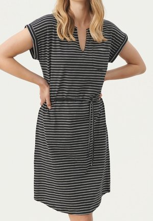 Femme portant une robe à manches courtes rayée horizontalement en noir et blanc, avec une ceinture à la taille et une petite poche poitrine, bras pliés avec les mains sur les hanches.