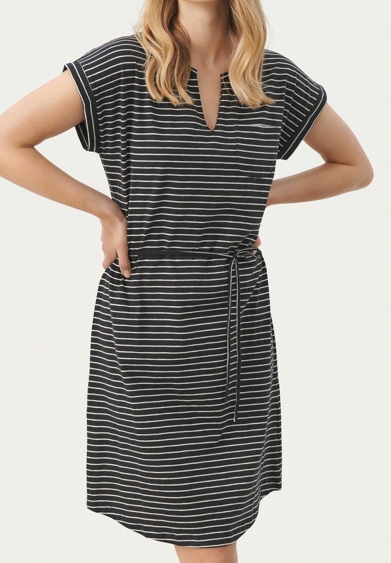 Femme portant une robe à manches courtes rayée horizontalement en noir et blanc, avec une ceinture à la taille et une petite poche poitrine, bras pliés avec les mains sur les hanches.