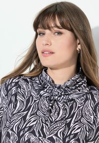 Blouse noir et blanc avec un motif de feuilles, tissu doux, col haut avec des détails de cordon, et accents de couture visibles.