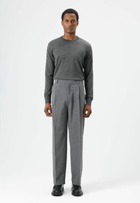 Chemise grise à manches longues associée à un pantalon gris plissé. La tenue présente une coupe décontractée avec une texture lisse et des détails subtils. Des chaussures noires complètent le look.