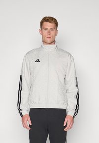 Giubbotto bomber grigio chiaro con un sottile motivo a rilievo, logo Adidas nero e strisce nere sulle maniche, completato da un bordo elastico.