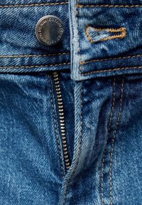 Jean en denim avec un bouton métallique argenté et une fermeture éclair visible, avec des coutures marron contrastantes sur un tissu bleu texturé.