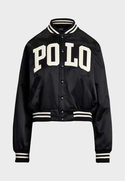 Must mustang bomber-jakk musta satiniga, millel on valged "POLO" kirjutised. Omab triibulisi soonikpuud ja vöökohta, nuppe ja küljetaskuid.