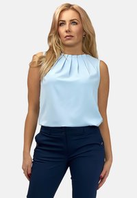 Jasnoniebieska bluzka bez rękawów z plisowanym dekoltem, w połączeniu z granatowymi spodniami w klasycznym kroju, które mają zasuwaną kieszeń i zapięcie na guzik.