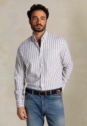 Homme portant une chemise boutonnée à rayures verticales blanches et grises, rentrée dans un jean bleu avec une ceinture tissée noire, sur un fond neutre.