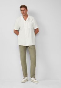 Chemise blanche à manches courtes avec poche, associée à un pantalon vert clair. Le modèle porte des baskets blanches avec des accents beiges. Design simple, texture lisse.