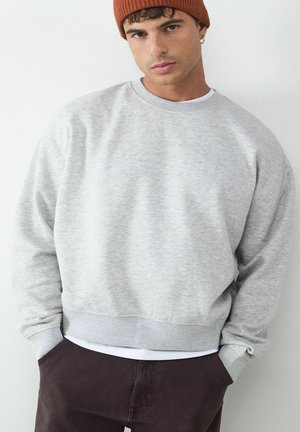 Lichtgrijze sweatshirt met een ronde hals, ribbelboorden en onderrand. Draag het over een wit shirt, gecombineerd met donkerbruine broeken en een roestkleurige beanie.