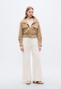 Κοντό μπεζ σακάκι με δύο μπροστινές τσέπες, σε συνδυασμό με λευκό παντελόνι καμπάνα και καφέ παπούτσια. Ομαλή υφή και μεταλλικά διακοσμητικά στοιχεία.