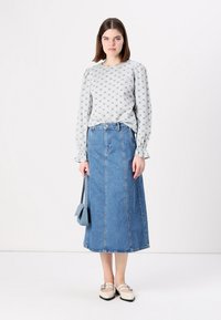 Blusa cinza com padrão e mangas bufantes, combinada com uma saia de ganga de cintura alta, sapatos bejes e uma mala azul pequena.
