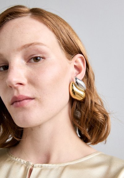 Boucles d'oreilles en métal doré et argenté au design courbé et abstrait. Des surfaces lisses et brillantes avec des couleurs et des formes contrastées créent une apparence semblable à un géode.