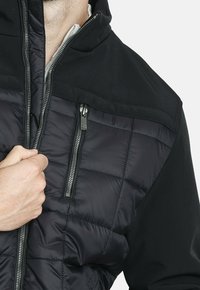 La mano de un hombre cerrando una chaqueta negra acolchada con un bolsillo en el pecho con cremallera vertical, contra un fondo claro y liso.