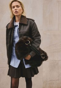 Jachetă maro din piele cu guler de blană, cămașă bleu deschis cu nasturi, fustă neagră plisată, ținând o geantă mare de blană maro cu curea.