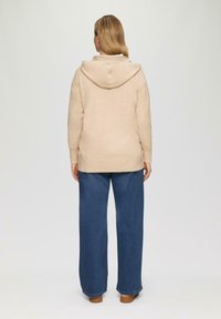 Beige Kapuzenpulli mit weicher Textur, lockerer Passform und gerippten Bündchen, kombiniert mit weiten blauen Jeans.