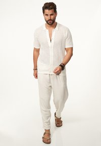 Weißes gestricktes Shirt mit kurzen Ärmeln und Knopfleiste, kombiniert mit hellbeigen, locker sitzenden Hosen und braunen Sandalen.