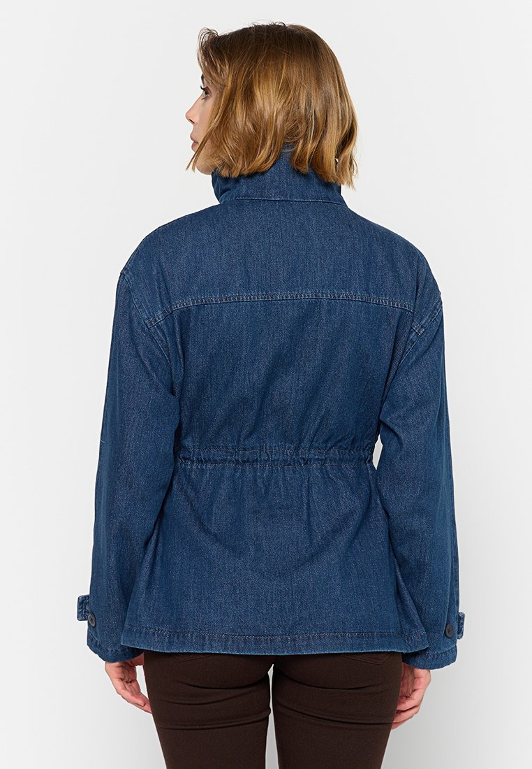 Mujer con cabello castaño corto que lleva una chaqueta de mezclilla azul oscuro y pantalones oscuros, de espaldas contra un fondo blanco liso.