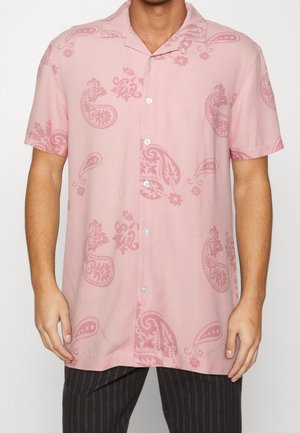 Mand iført en lyserød kortærmet skjorte med knapper og mørkere pink paisley-mønstre samt mørke stribede bukser.