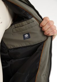 Interno giacca verde oliva con foderatura nera, dotato di tasca a toppa e etichetta con logo. Zip a vista con profili marroni.
