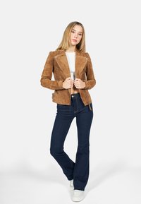 Bruine suede motorjack met zilverkleurige accenten, aansluitend ontwerp, asymmetrische rits en zijgespen, gecombineerd met donkerblauwe flared jeans.