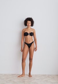Black bandeau bikini with a gathered design and center knot detail. High-cut bikini bottom with minimal coverage and accent hardware on the sides.