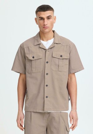 Junger Mann trägt beige kurzärmliges Hemd mit Brusttaschen über weißem T-Shirt, kombiniert mit passenden beigen Hosen.