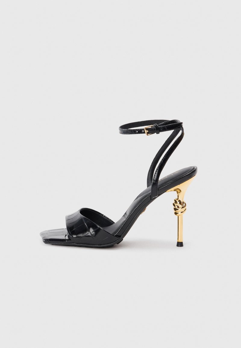 Sandales à talons hauts noires avec une finition texture crocodile. Présente une fine bride à la cheville et un accent en métal doré torsadé sur le talon. Bout carré.
