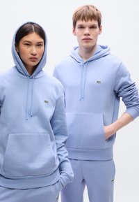 Ανοιχτό μπλε φούτερ με κουκούλα και αντίστοιχα παντελόνια, κατασκευασμένα από μαλακό ύφασμα, με μπροστινή τσέπη και μικρές πράσινες λογότυπες λεπτομέρειες.