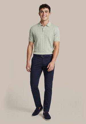 Man draagt lichtgroene gebreide korte mouwen polo, marineblauwe slim-fit broek met riem en marineblauwe instapschoenen, staand en glimlachend tegen een beige achtergrond.