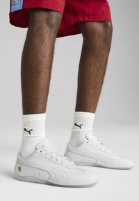 Zapatillas de cuero blanco con diseño perforado, con un logo amarillo de Ferrari, combinadas con calcetines blancos acanalados que exhiben un logo negro de Puma.