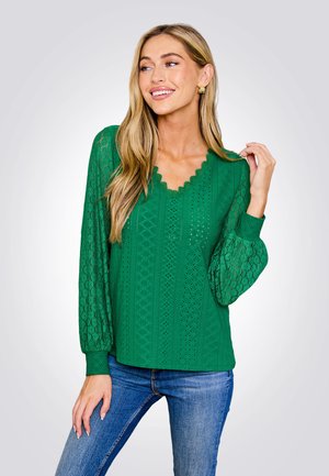 Groene kanten blouse met een gegolfde V-hals, lange ballonmouwen en textuurpatronen. Gecombineerd met blauwe jeans.