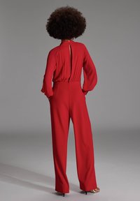 Rode jumpsuit met lange, vloeiende mouwen en een hoge halslijn. Heeft een sleutelgatrug en een getailleerde taille, uitlopend in wijde broekspijpen.