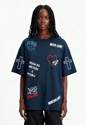 Vrouw die een oversized donkerblauw T-shirt draagt met haai, hart, cherubijnen, kruisen en tekst inclusief "Dream big," "Together" en "With love."