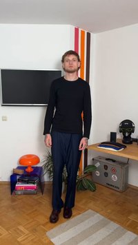 Sort langærmet skjorte, marineblå bukser og brune lædersko. Mønstrede detaljer på væggene, orange lampe og stablede tøj på et bord.