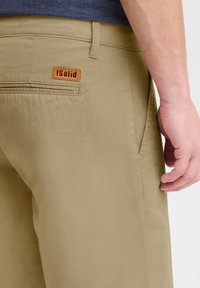 Beige broek van zachte stof, met een tan leren label met de tekst "Solid" op de achterzak en een verborgen ritszakdetail.