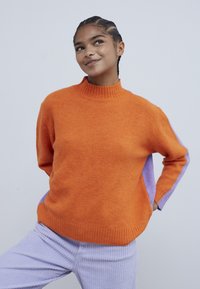 Jeune personne souriante, portant un pull orange avec des manches violettes et un pantalon en velours côtelé violet clair, debout devant un fond uni.