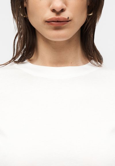 Νεαρό άτομο με σκούρα μαλλιά μήκους μέχρι τους ώμους φορά ένα απλό λευκό πουκάμισο, δείχνοντας τα χείλη και ένα έντονο σκουρόχρωμο σπυράκι πάνω από το πάνω χείλι.