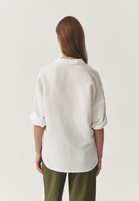 Camicia bianca in lino a vestibilità relax con maniche arrotolate e colletto, caratterizzata da un orlo curvo e una texture morbida. Abbinata a pantaloni verde oliva.