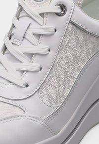 Κοντινό πλάνο ενός λευκού αθλητικού παπουτσιού με επάνω μέρος από υφασμάτινη υφή, λευκά κορδόνια, μεταλλικές γυαλιστερές οπές και λεπτομέρειες στη ραμμένη σόλα.