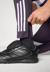 Zapatilla atlética negra con parte superior texturizada y detalles a rayas, combinada con pantalones deportivos grises con rayas blancas laterales y puños con cremallera.