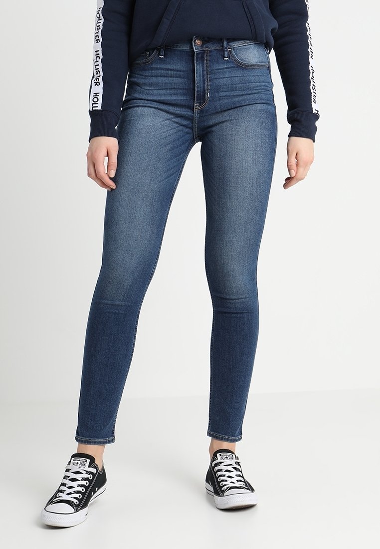 Jeans in denim blu slim-fit con lavaggio medio, leggera scoloritura e design a cinque tasche, abbinati a sneakers in tela bianca e nera.