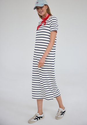 Kobieta w białej i granatowej sukience w paski do połowy łydki, jasnobłękitnej czapce, czerwonej bandanie i białych trampkach stoi na jednolitym tle.