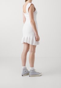 Robe blanche en tricot avec un ourlet évasé et des bretelles festonnées, associée à des baskets montantes grises et des chaussettes blanches. Design simple et texturé.